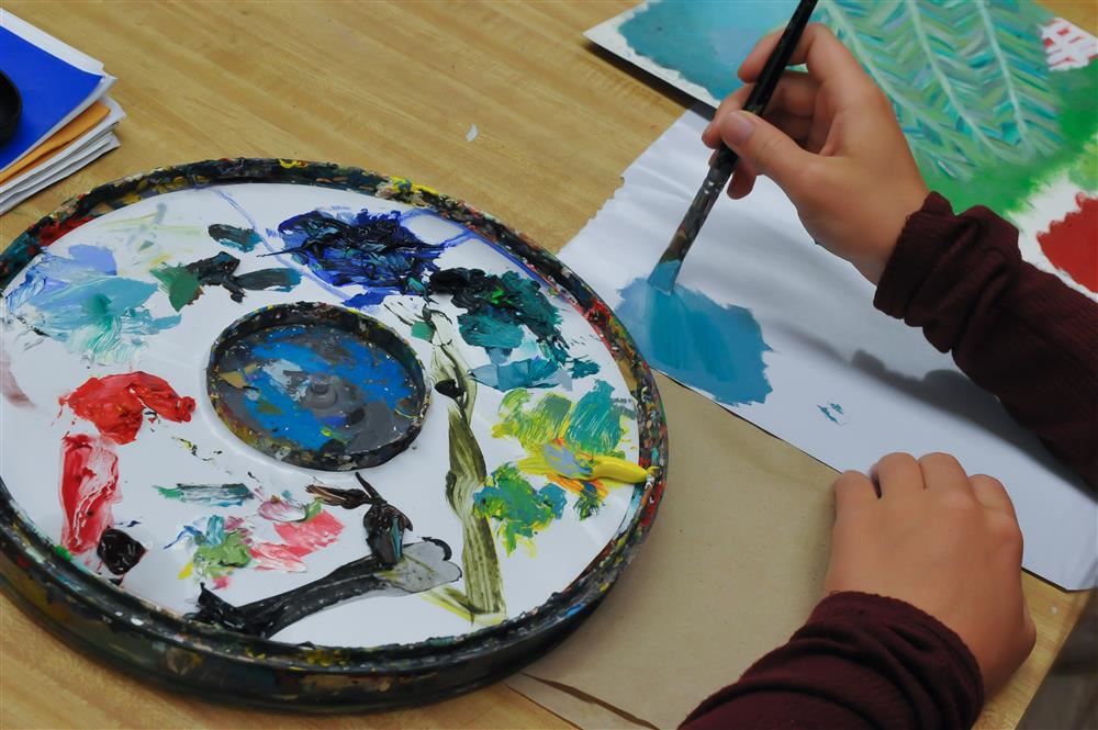 Hand painting with a brush on paper with a palette of paints beside him.