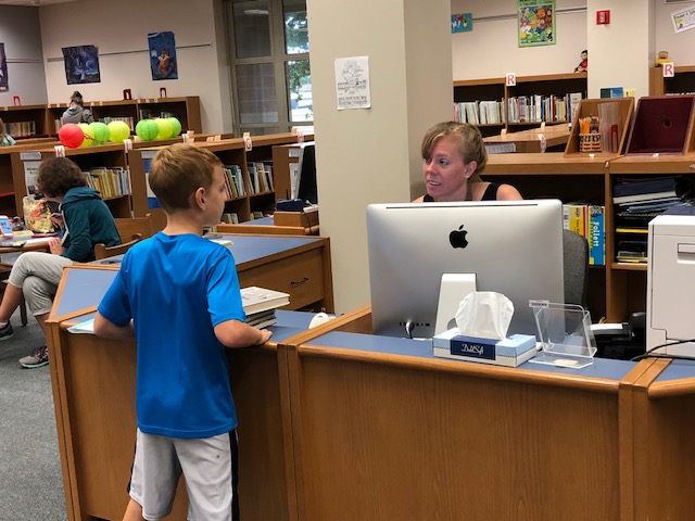 A student holding a book with a librarian behind their desk. A student holding a book with a librarian behind their desk.